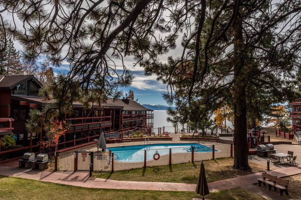 Outdoor fenced pool framed by pine branches, with lodge buildings and Lake Tahoe beyond