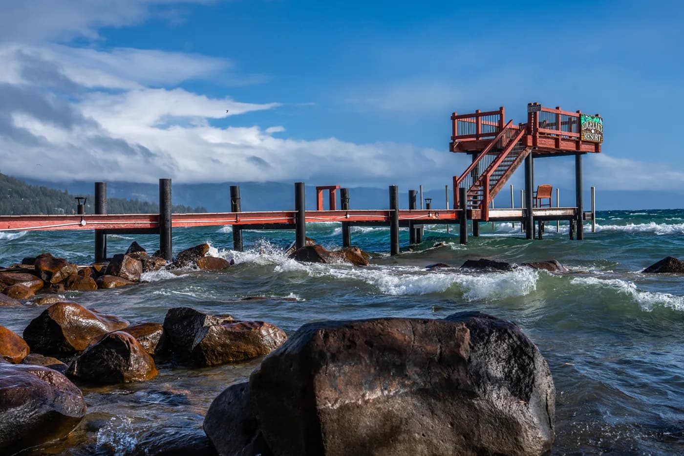 Wooden pier with upper deck overlooking a rocky shoreline and vibrant blue lake.