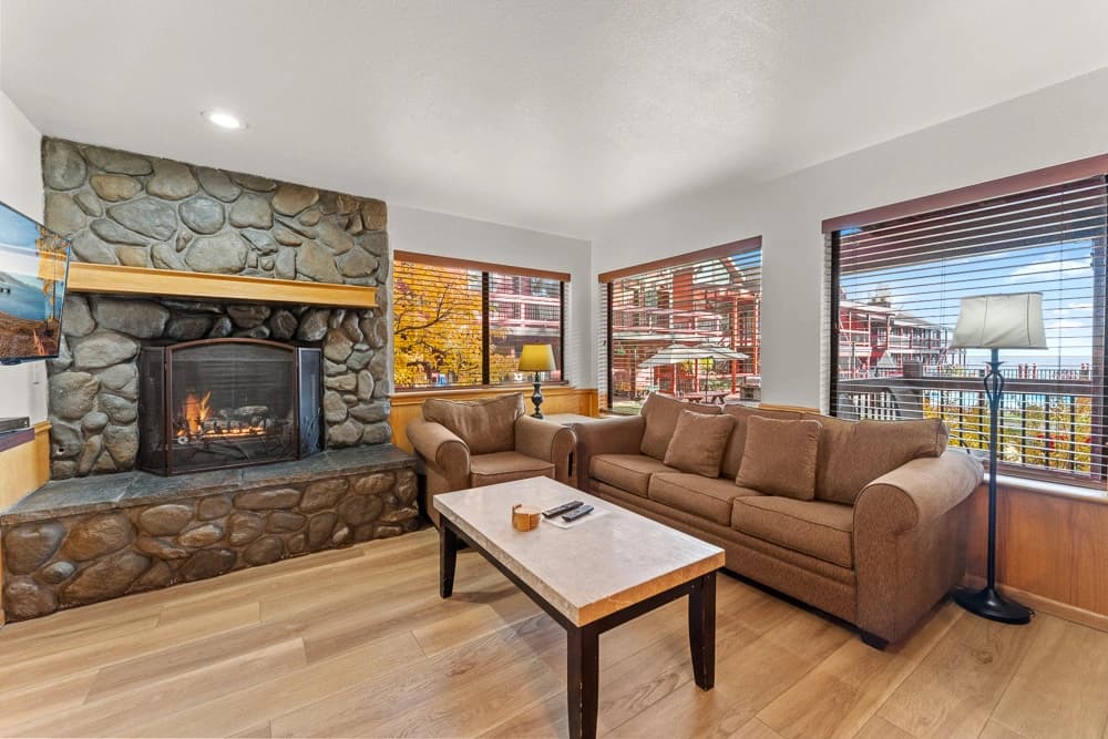 Cozy living room with stone fireplace, plush sofas, and large windows with scenic forest view.